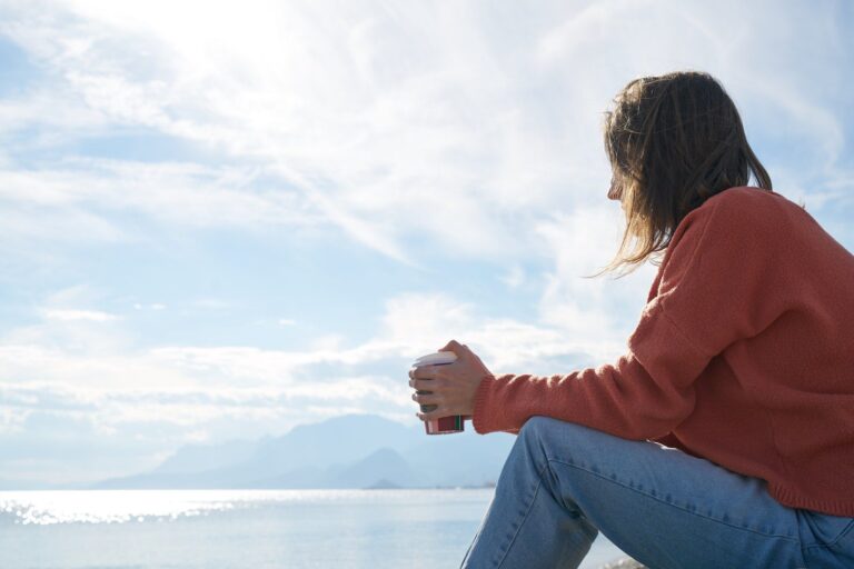 A woman in a rust-colored sweater sits by a calm sea holding a coffee cup, gazing toward distant mountains in quiet reflection — evoking emotional self-awareness and self-regulation.